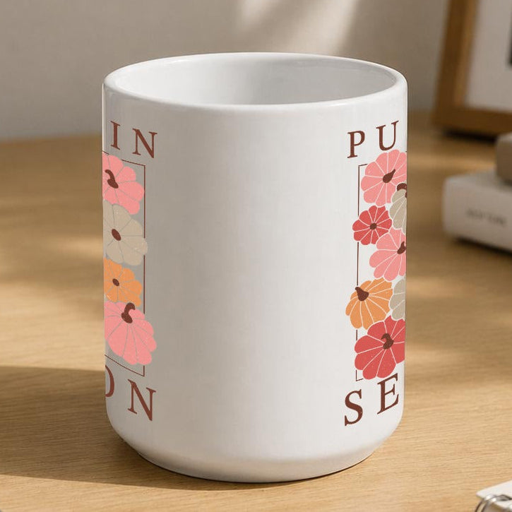 A white ceramic mug with a pink and orange floral design and partial text, sitting on a wooden desk in a styled home office setting with a laptop, notebook, and plant.