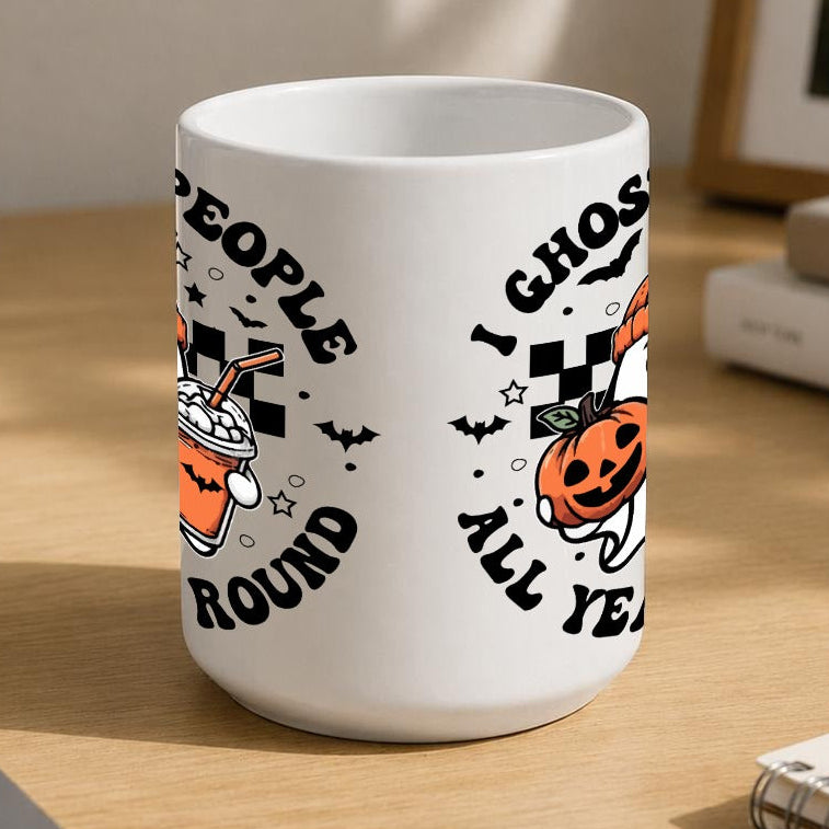 A white ceramic mug with a Halloween-themed graphic print sits on a wooden desk in a styled home office setting. The mug features a cartoon ghost wearing an orange beanie and holding a jack-o'-lantern and an iced drink, surrounded by the text 'I GHOST PEOPLE ALL YEAR ROUND'.