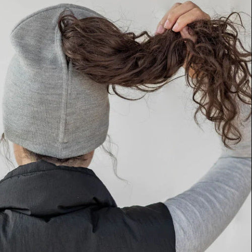 Person wearing a gray beanie and black vest, holding hair with a plain background