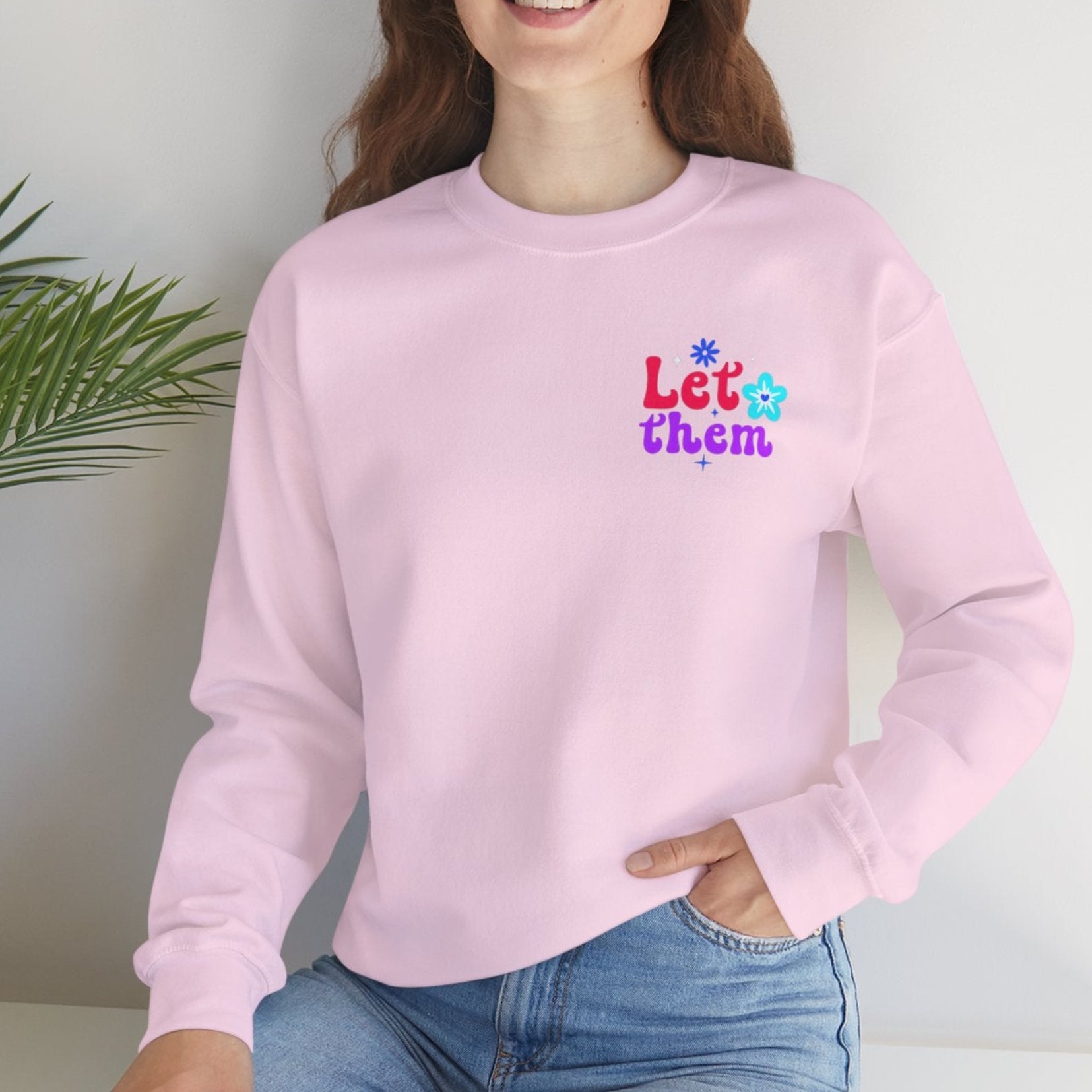Person wearing a pink sweatshirt with colorful text, sitting on a white surface with a plant in the background.