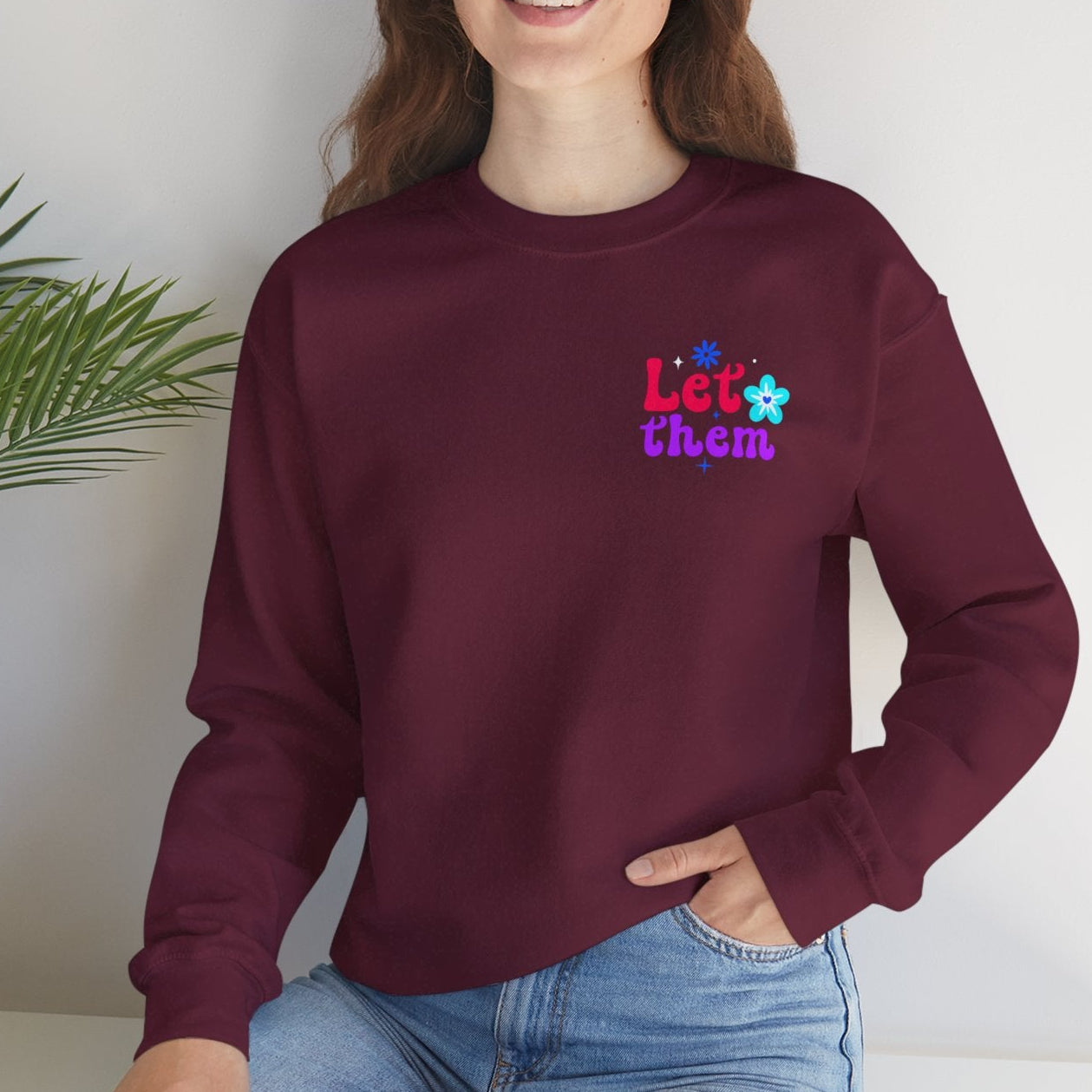 Person wearing a maroon sweatshirt with colorful text, sitting on a white surface with a plant in the background.