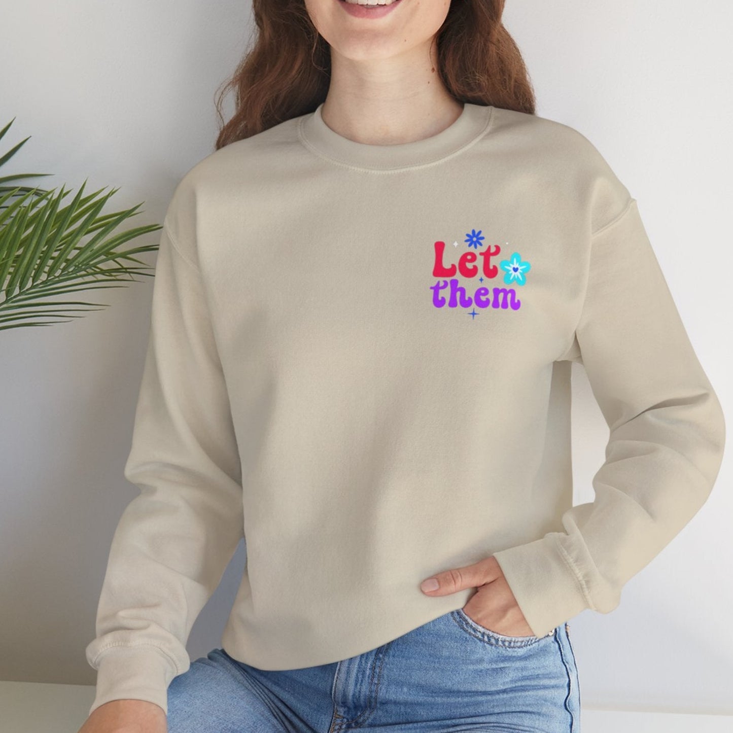Woman wearing a sand sweatshirt with colorful text sitting on a white surface with a plant in the background