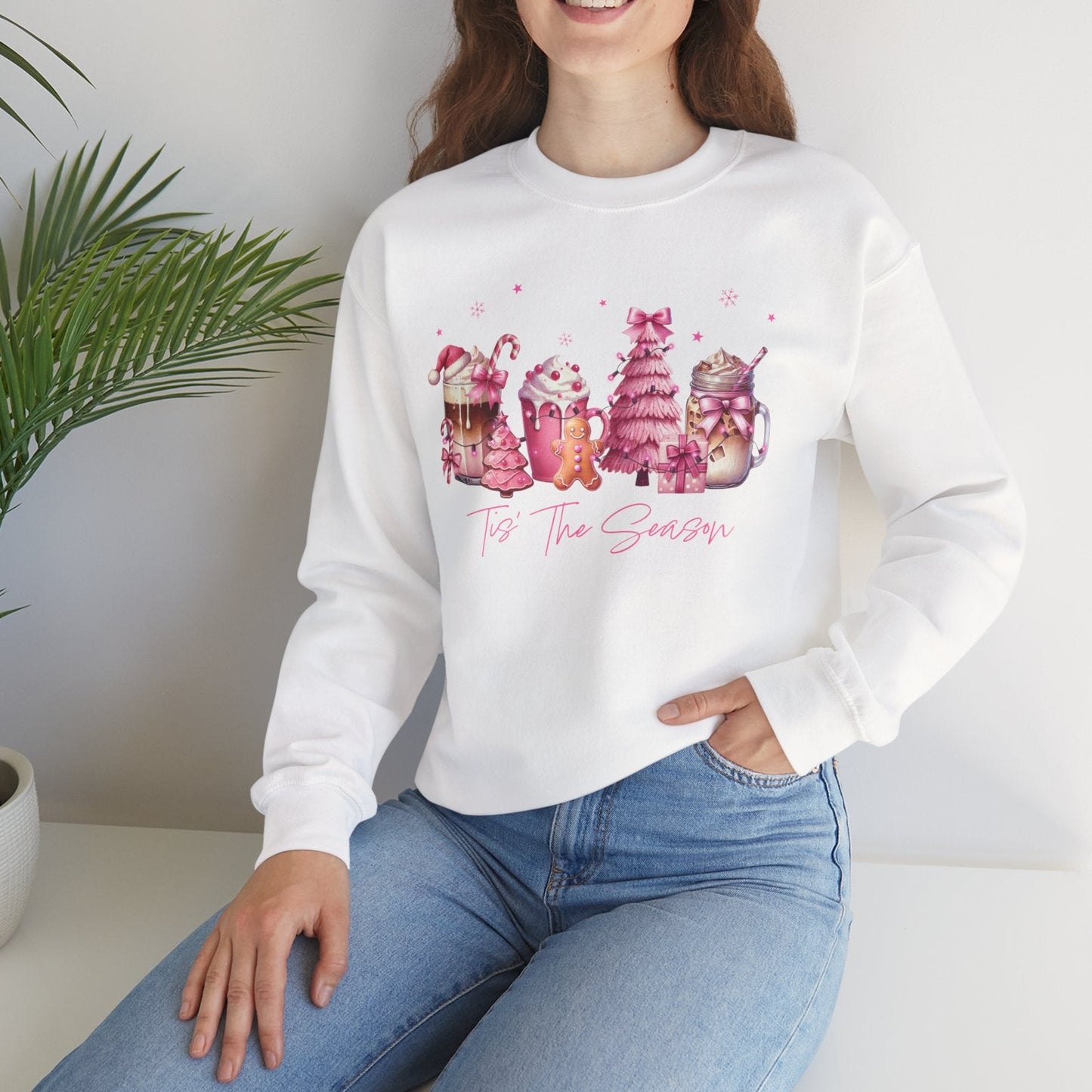 Woman wearing a white sweatshirt with colorful graphics and text, sitting in a room with a plant.