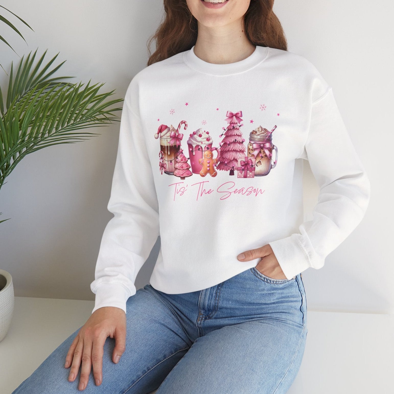 Woman wearing a white sweatshirt with colorful graphics and text, sitting in a room with a plant.