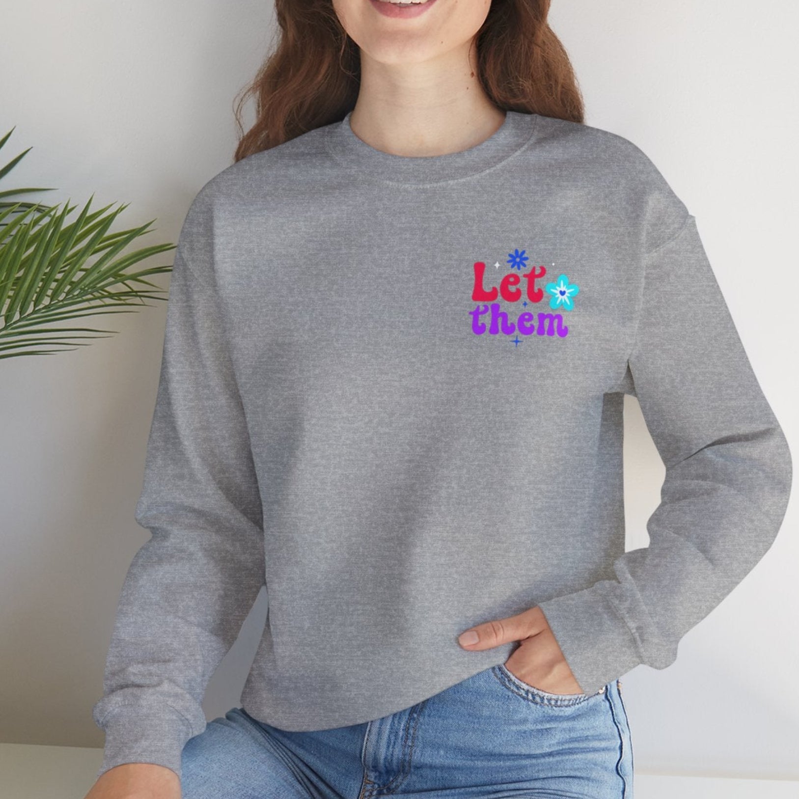 Woman wearing a gray sweatshirt with colorful text sitting on a white surface with a plant in the background