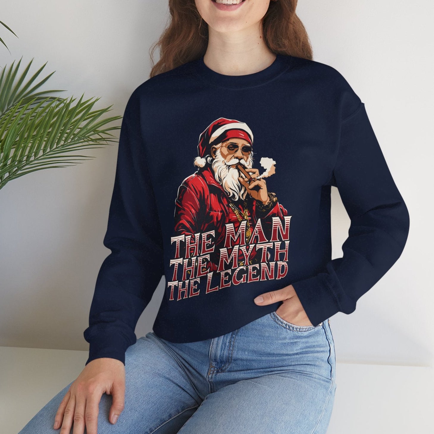 Person wearing a navy blue sweatshirt with a graphic design and text, sitting against a white wall with a plant.