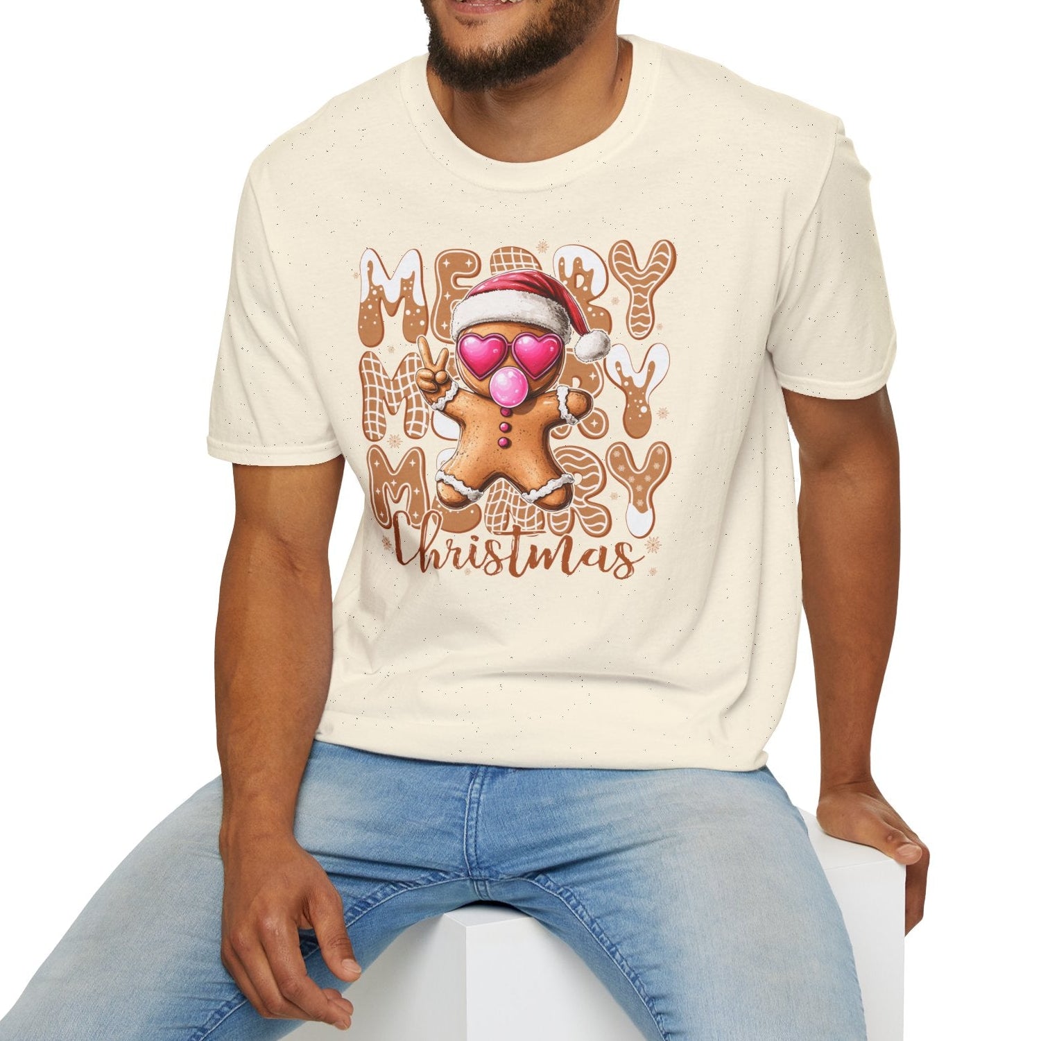Man wearing a cream t-shirt with a Christmas design, sitting on a white stool against a white background.