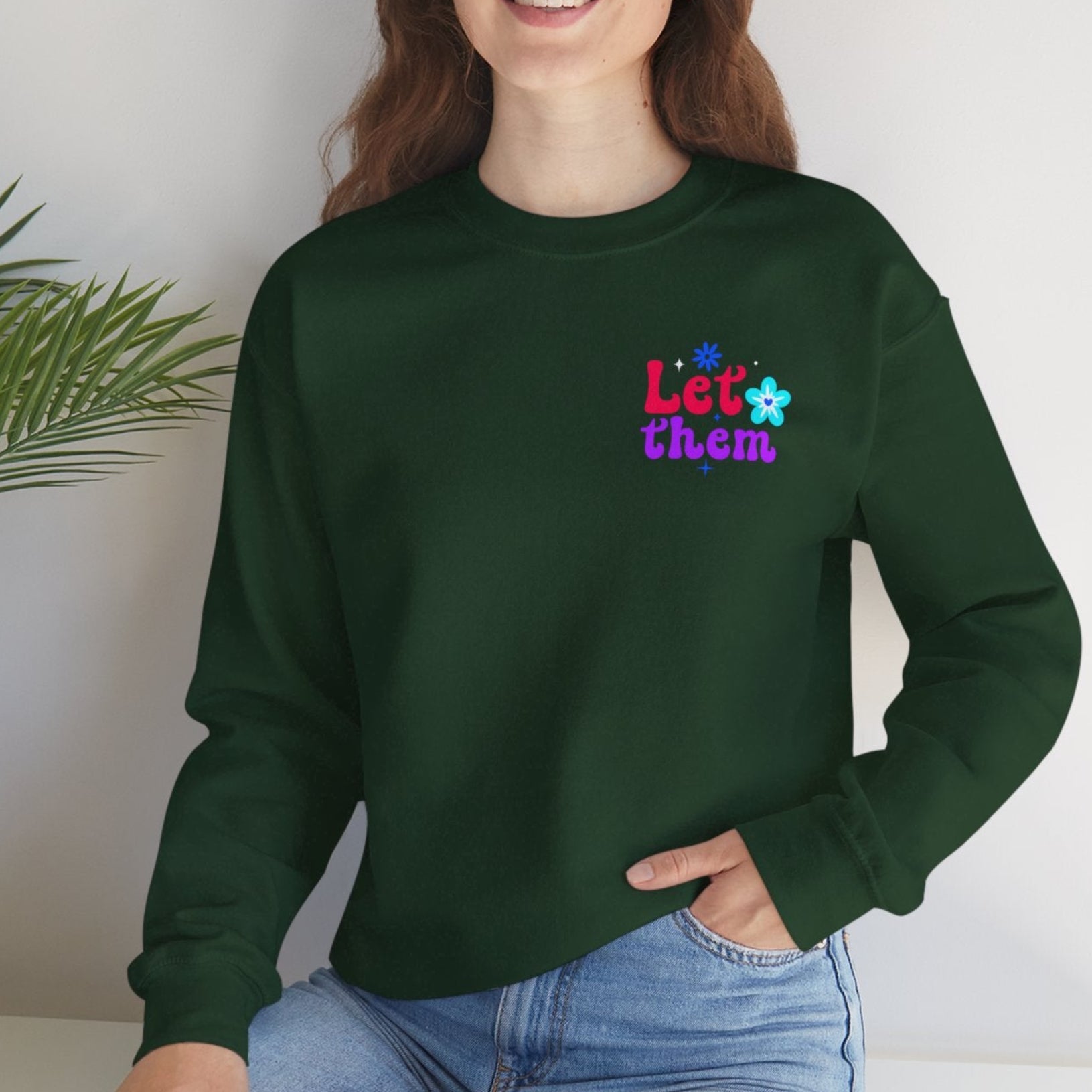 Person wearing a green sweatshirt with colorful text, sitting on a white surface with a plant in the background.