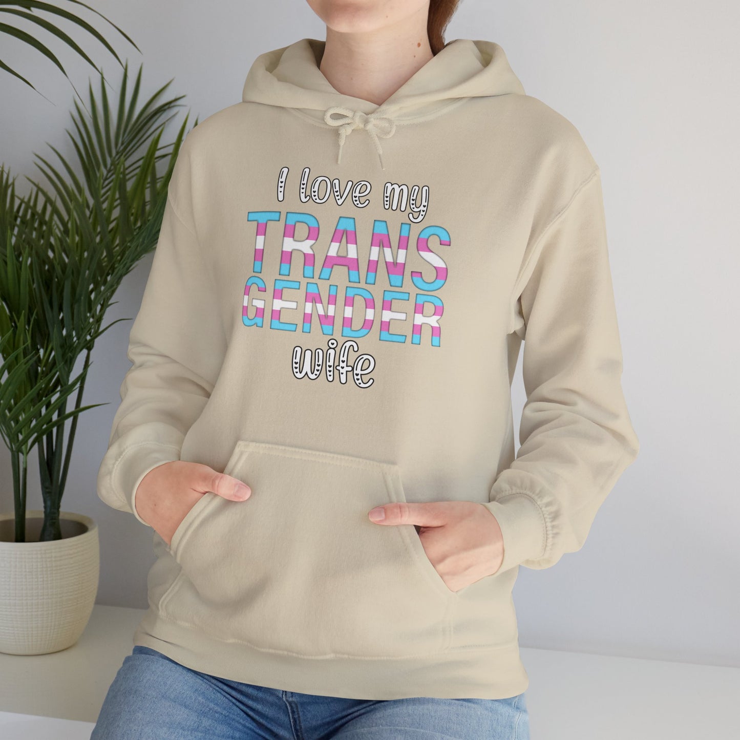 Person wearing a beige hoodie with colorful text 'I love my TRANS GENDER wife' in a home setting.