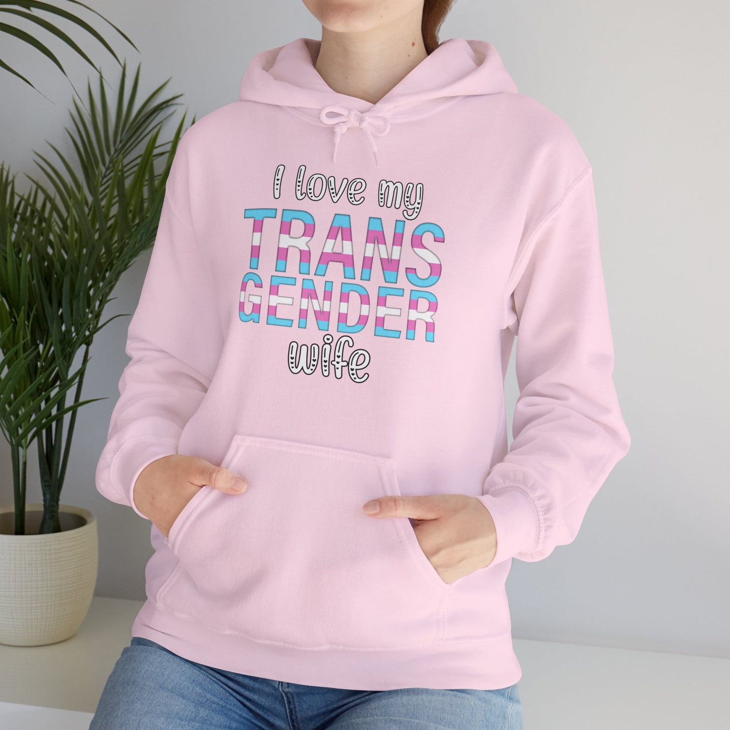 Person wearing a pink hoodie with text 'I love my TRANS GENDER wife' in a home setting.