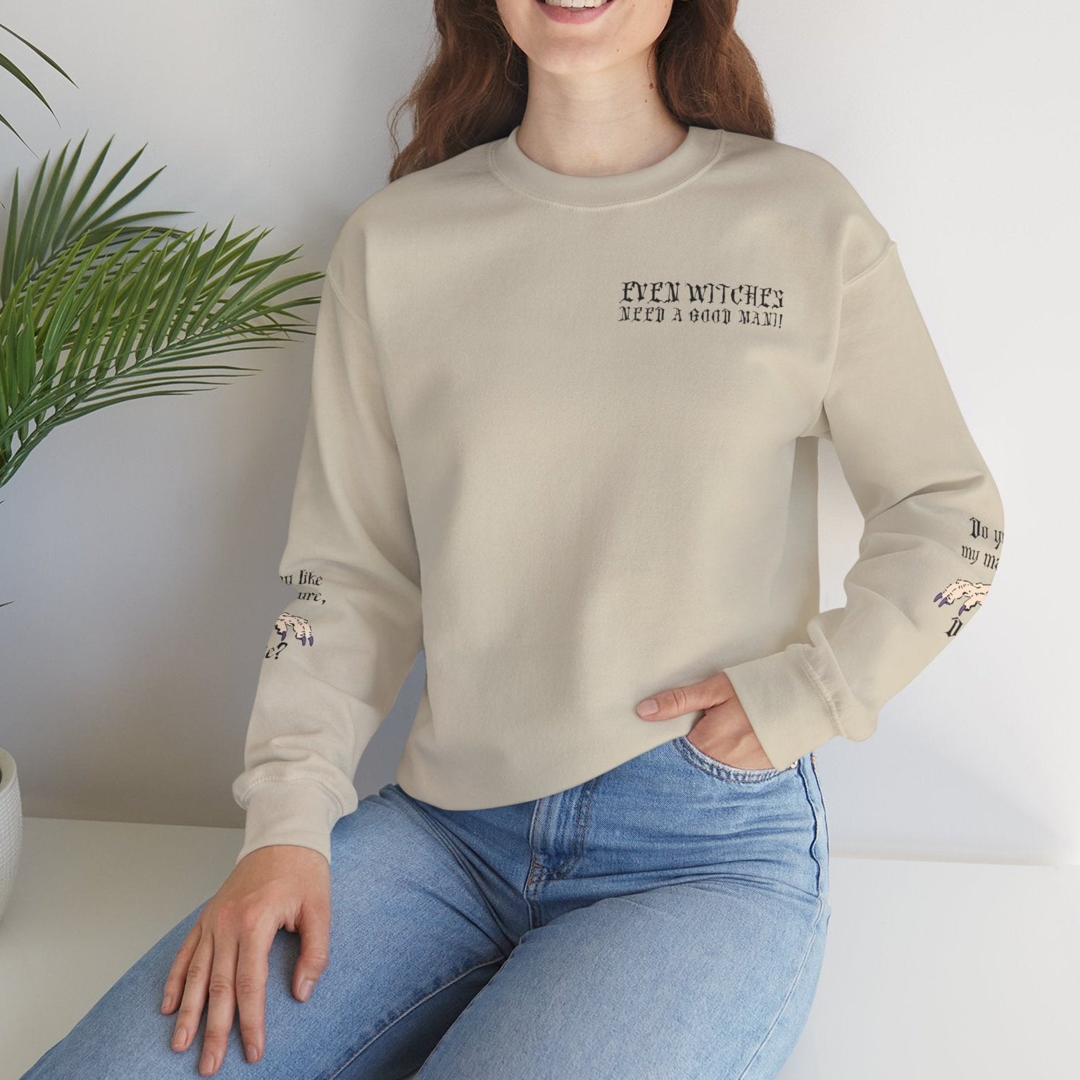 Woman wearing a beige sweatshirt with text and blue jeans sitting on a white surface with a plant in the background.