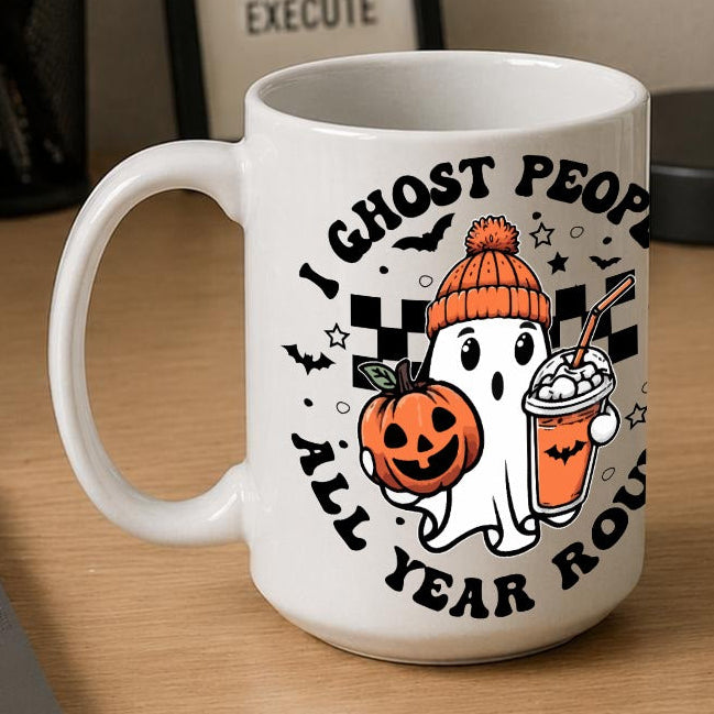 A white ceramic coffee mug with a Halloween graphic sits on a wooden desk next to a laptop, notebook, and framed sign. The mug features a cartoon ghost wearing an orange beanie and holding a jack-o'-lantern and an iced drink, surrounded by the text 'I GHOST PEOPLE ALL YEAR ROUND'.