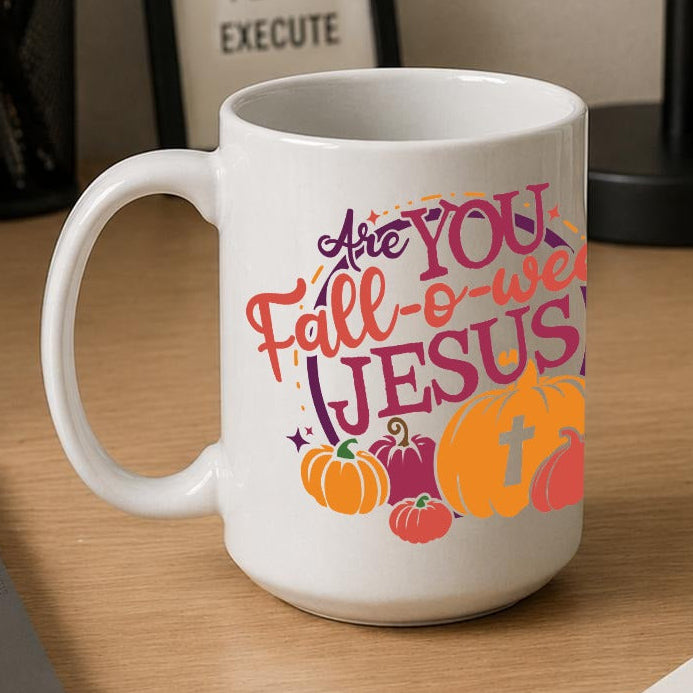 A white ceramic coffee mug with a colourful 'Are You Fall-o-ween JESUS' graphic sits on a wooden desk next to a laptop, notebook, and framed sign.