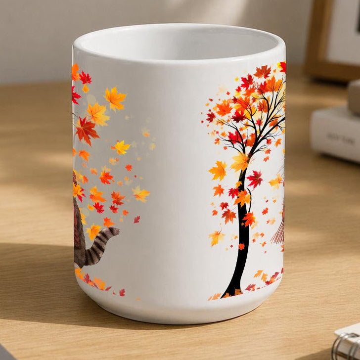 A white ceramic mug with a printed autumn tree and cat design, sitting on a wooden desk in a styled home office setting.