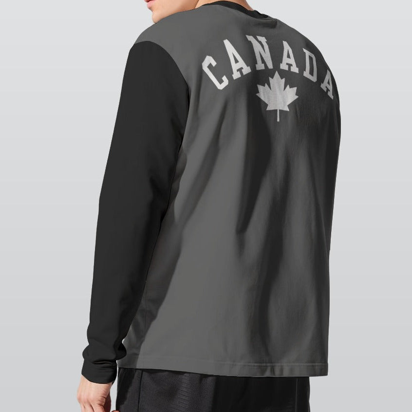 A man wearing a 2-toned dark gray and black long-sleeve athletic shirt with 'CANADA' and a maple leaf printed on the back, shown from a three-quarters angle against a light gray studio background.