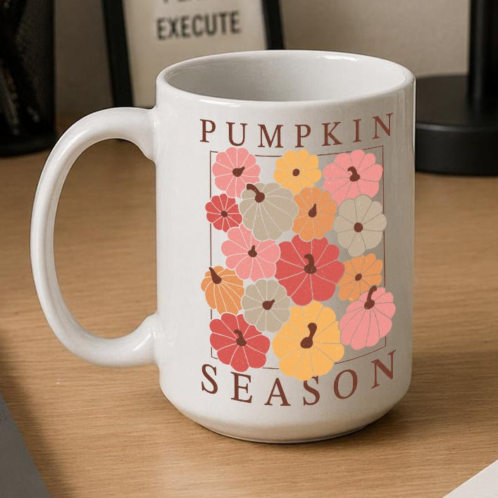 A white ceramic mug with a 'Pumpkin Season' floral graphic sits on a wooden desk next to a laptop, notebook, and framed sign.