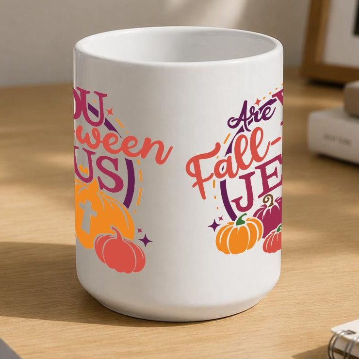 A white ceramic mug with a colourful autumn-themed graphic reading 'Are YOU Fall-O-ween JESUS' with pumpkins and fall leaves, placed on a wooden desk in a styled home office setting.