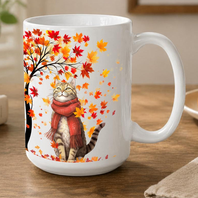 A white ceramic mug with a colourful illustration of a cat wearing a red scarf under an autumn tree with red and orange leaves, set on a wooden table in a cozy room.