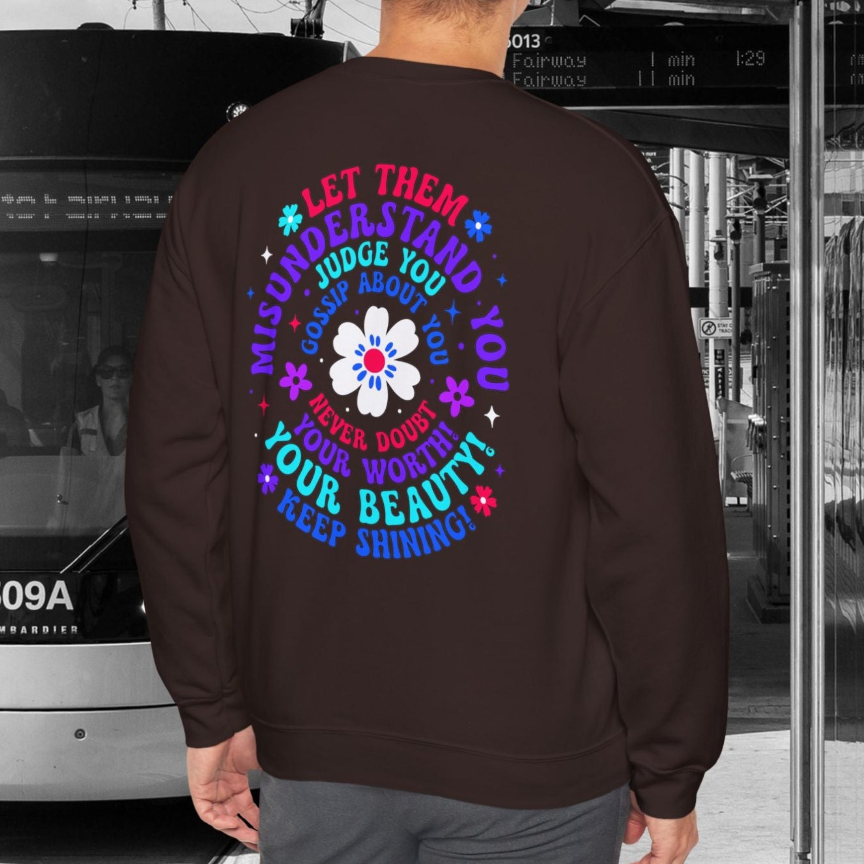 Person wearing a Chocolate brown sweatshirt with a colorful design standing next to a tram.
