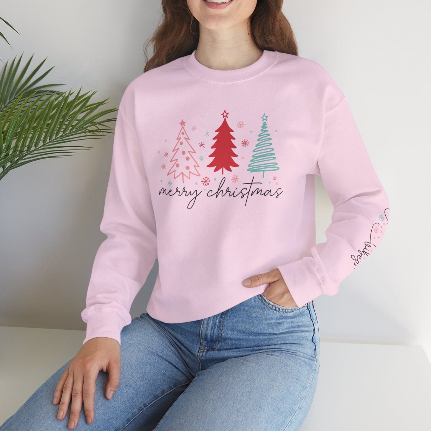 Person wearing a pink 'Merry Christmas' sweater with Christmas tree designs, sitting on a white surface with a plant in the background.