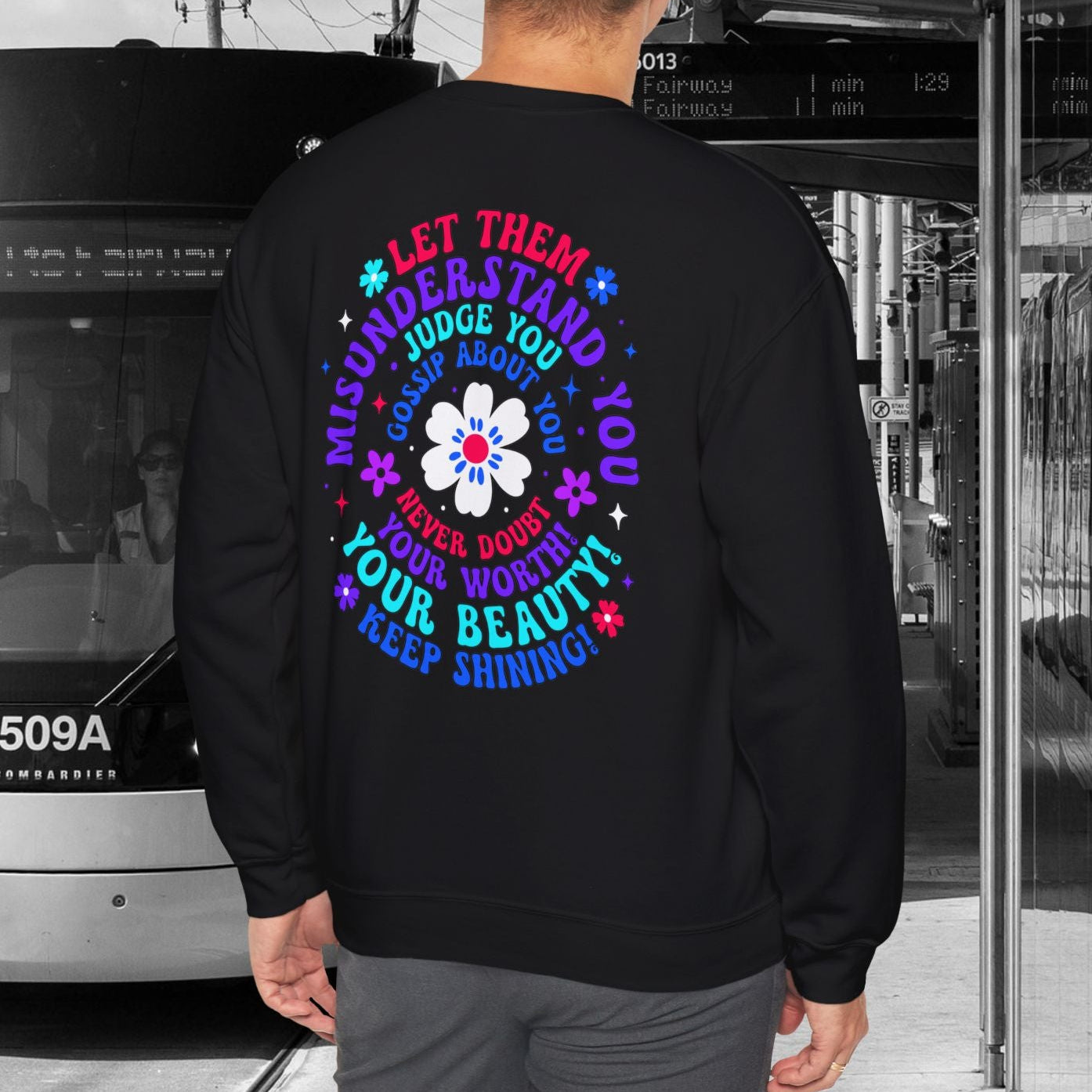 Person wearing a black sweatshirt with colorful text and a flower design, standing in front of a tram.