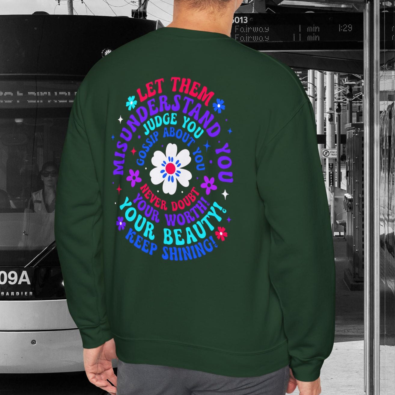 Person wearing a green sweatshirt with colorful text and design, standing in front of a tram.
