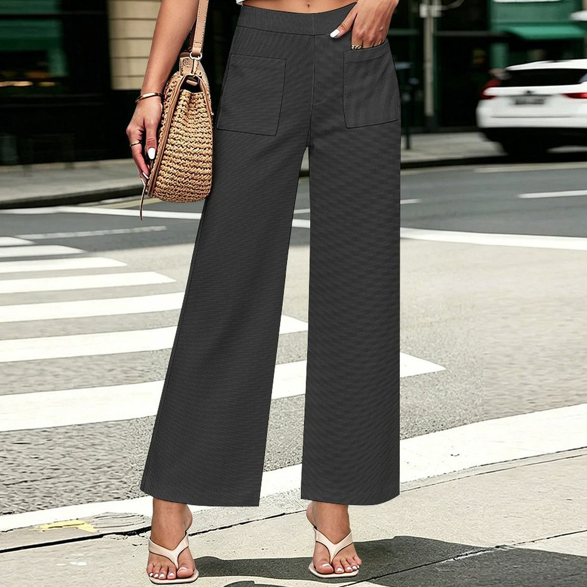 A person standing on a city crosswalk wearing dark charcoal faux corduroy wide-leg cropped pants, paired with a white crop top and beige flip-flops, while carrying a woven straw shoulder bag.