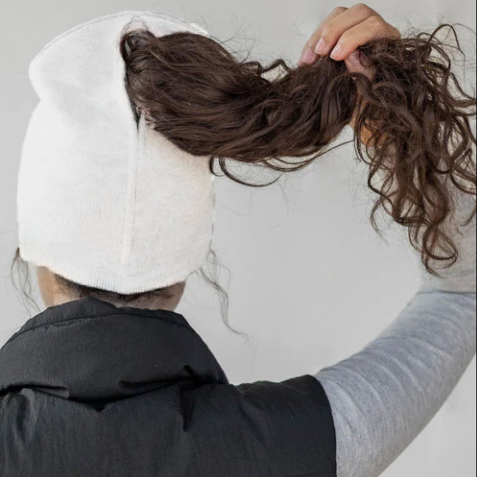 Person wearing a white beanie and black vest, holding their hair back against a plain background
