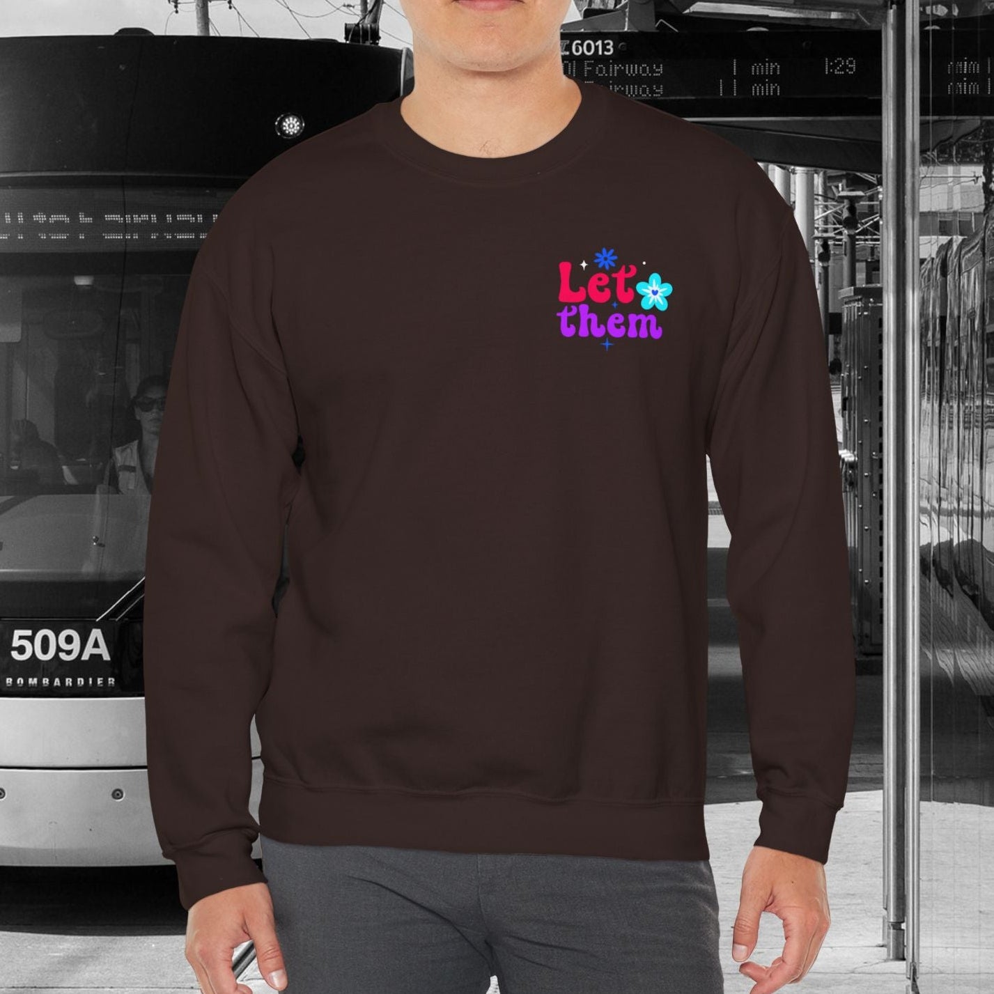 Man wearing a brown sweatshirt with colorful text standing in front of a tram.