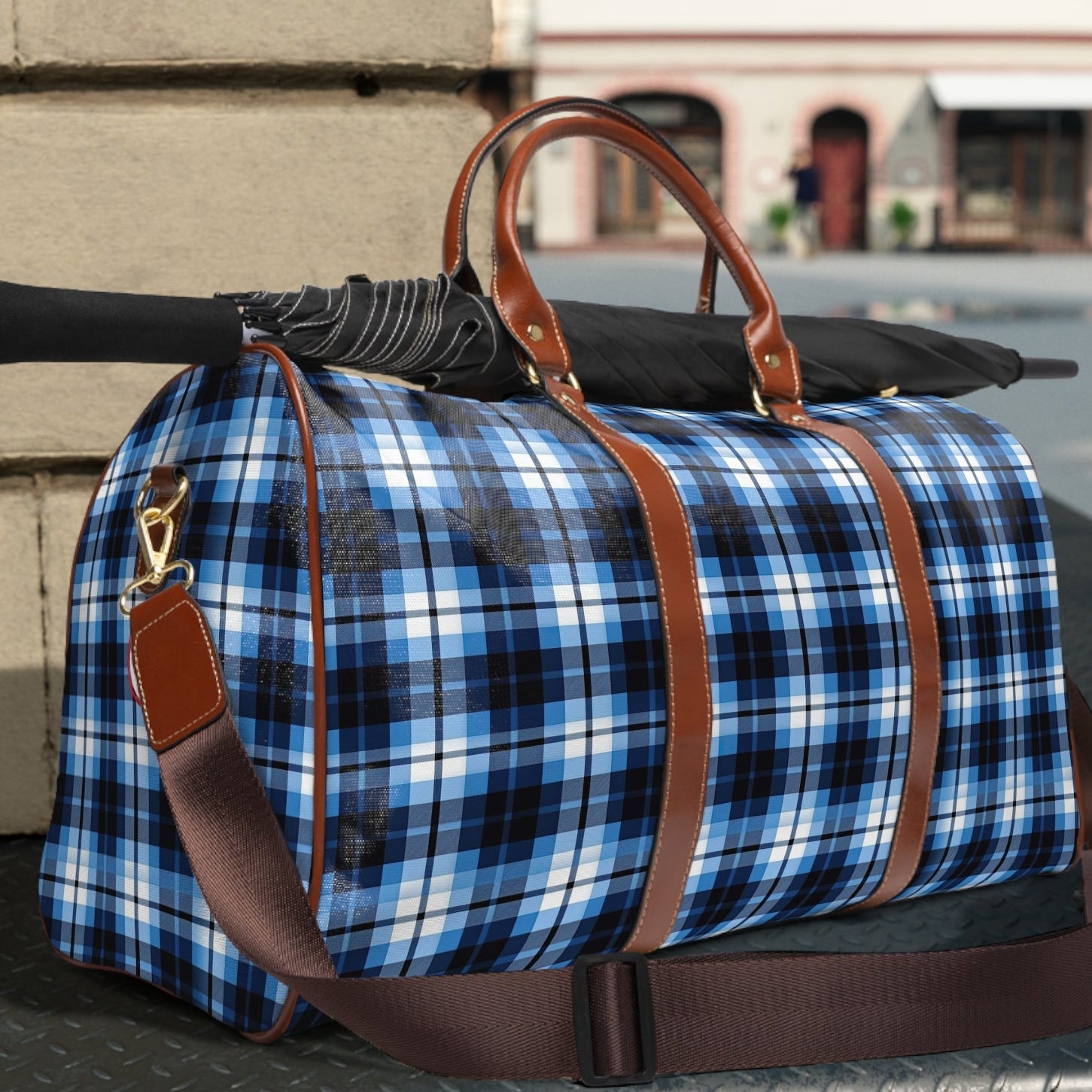 Blue plaid duffel bag with brown leather straps on a metal surface outdoors.