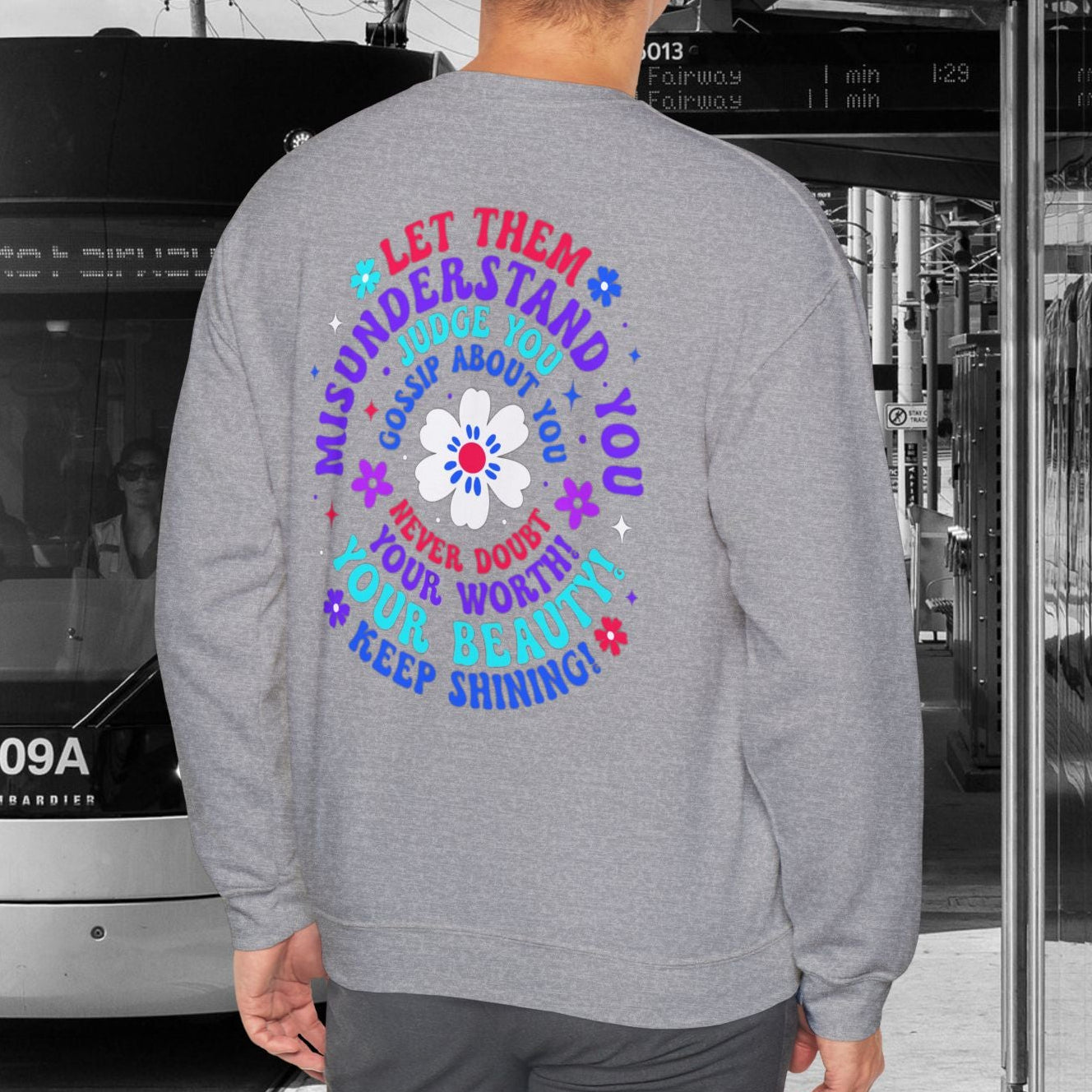 Person wearing a gray sweatshirt with colorful text and design, standing in front of a tram.