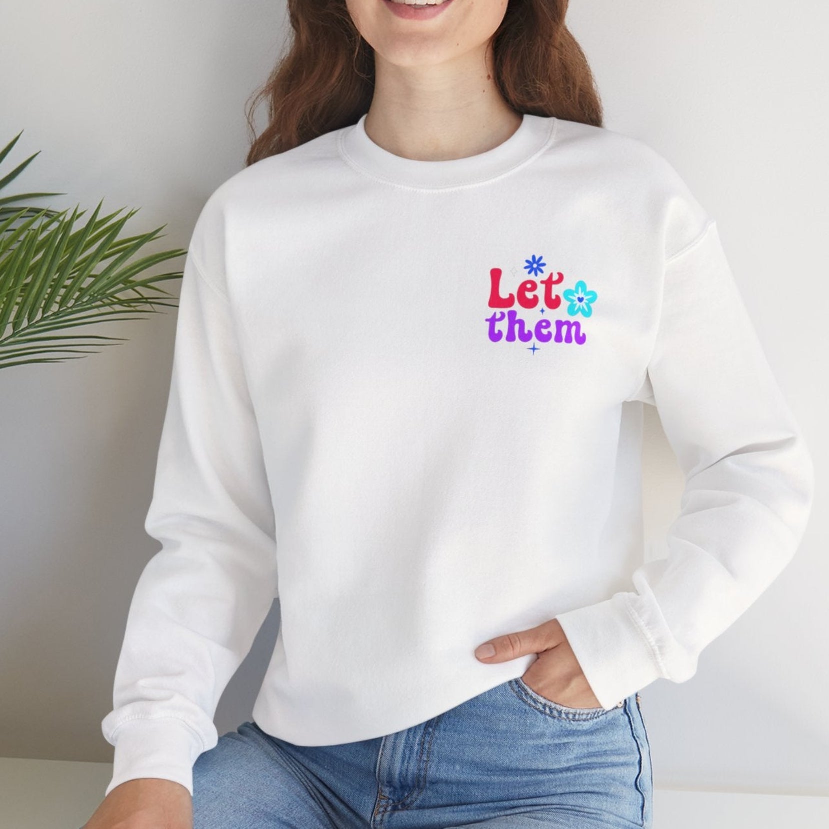 Person wearing a white sweatshirt with colorful text sitting on a white surface with a plant in the background