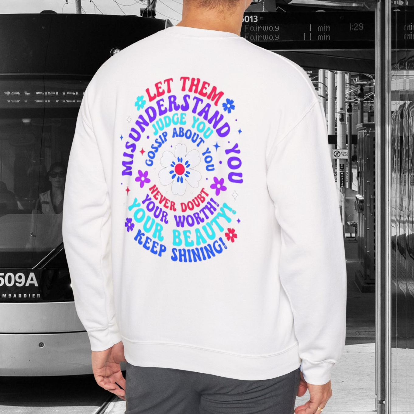 Person wearing a white sweatshirt with colorful text design, standing in front of a tram.