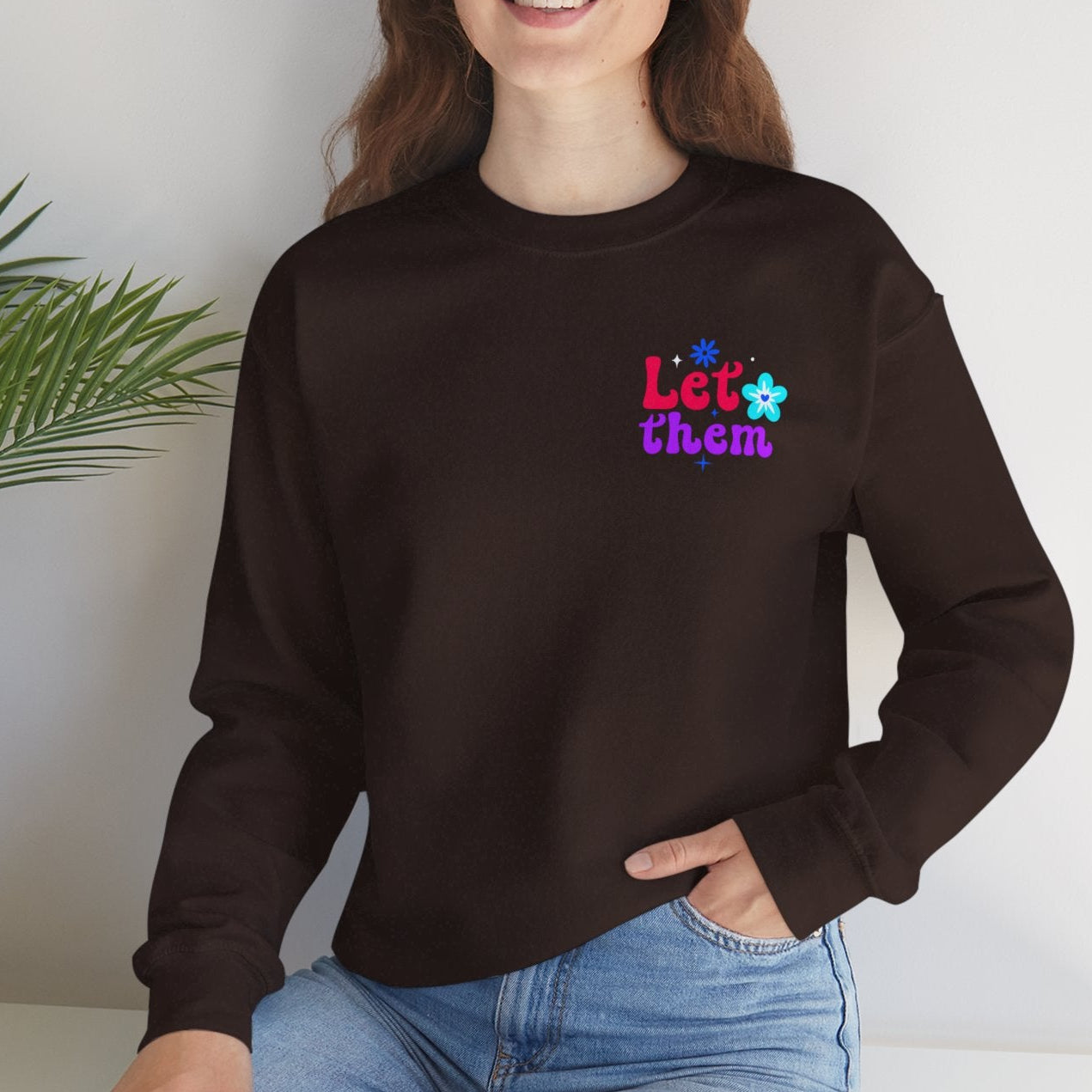 Person wearing a chocolate brown sweatshirt with colorful text, sitting on a white surface with a plant in the background.