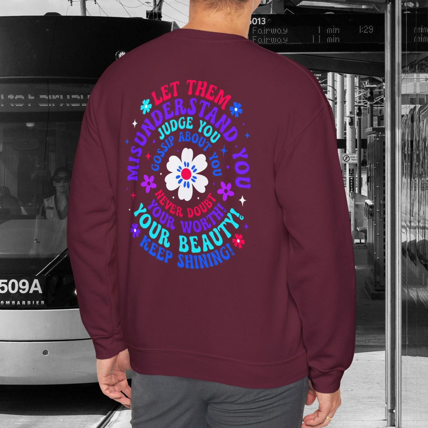 Person wearing a maroon sweatshirt with colorful text and graphics, standing in front of a tram.