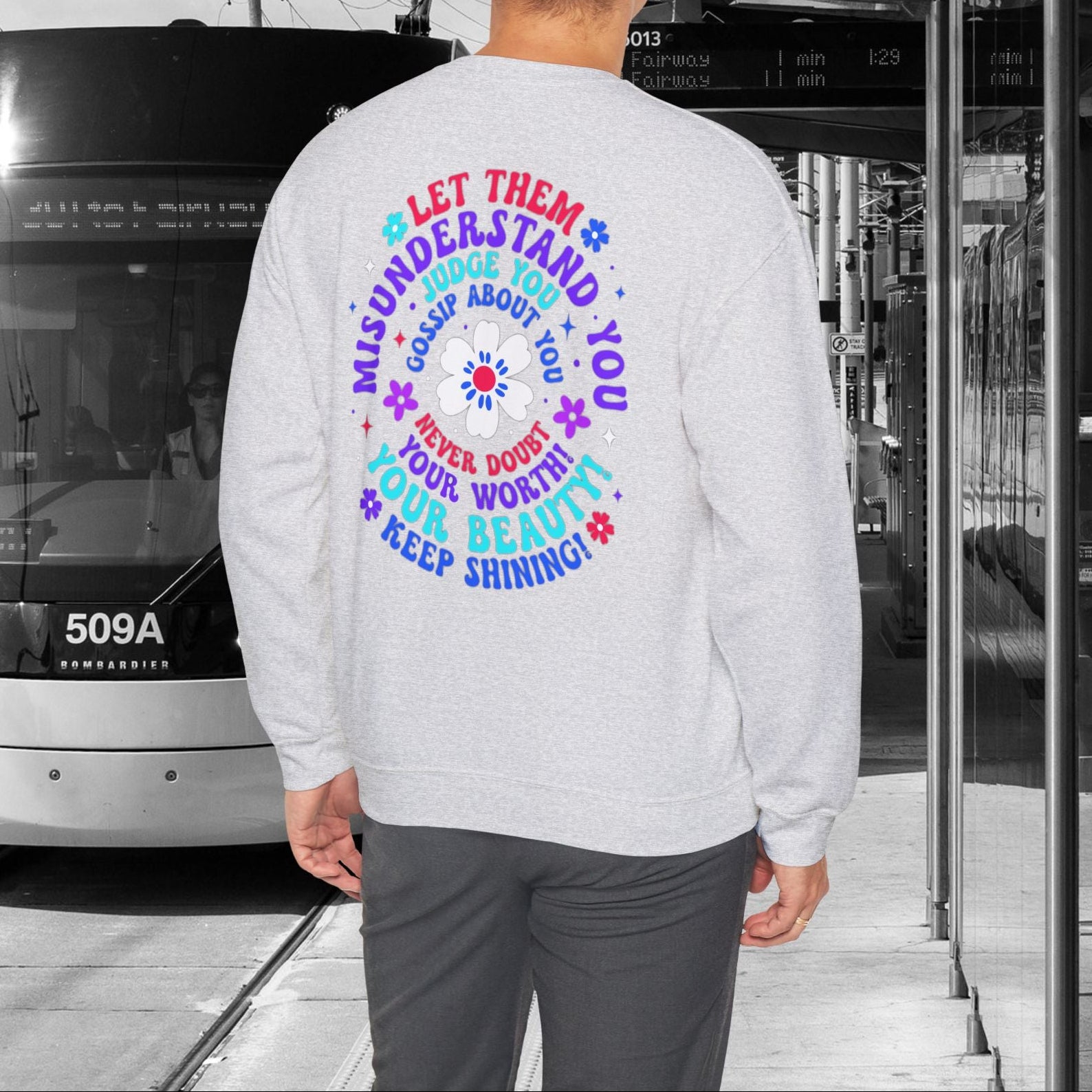 Person wearing an ash gray sweatshirt with a colorful message standing next to a tram.