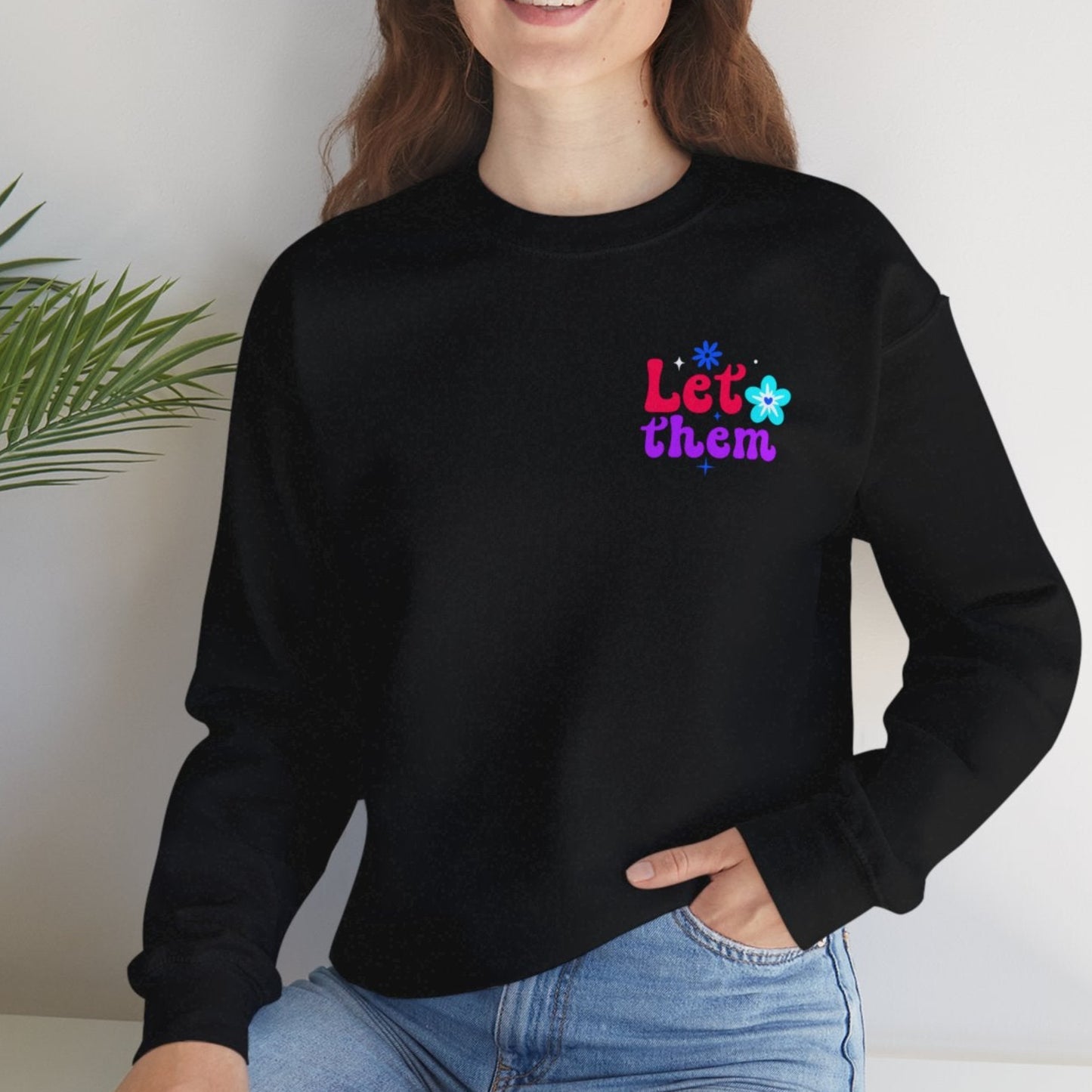 Person wearing a black sweatshirt with colorful text, sitting on a white surface with a plant in the background.