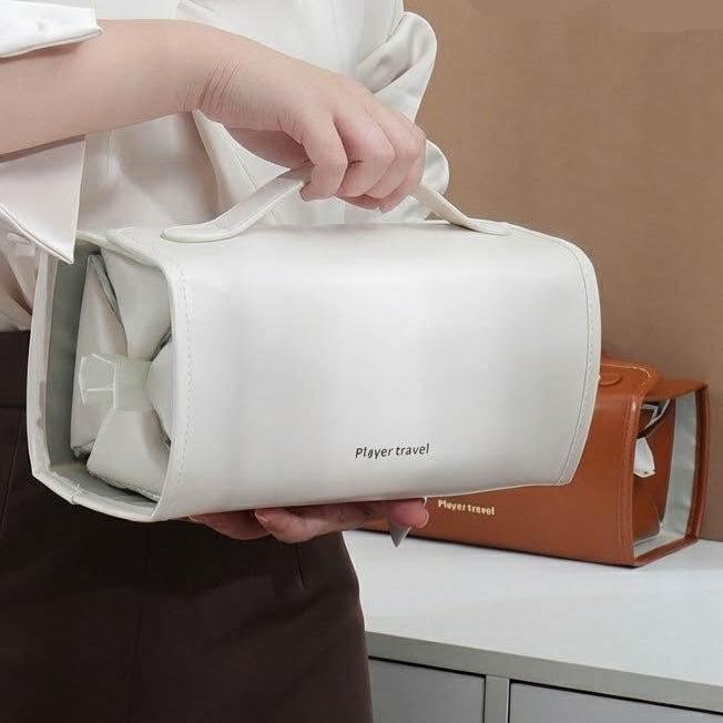 A person in a white blouse and brown skirt holds a white double-handle travel toiletry bag with a small printed logo. The scene is indoors with a beige wall, white vanity, and a small orange bag in the background.