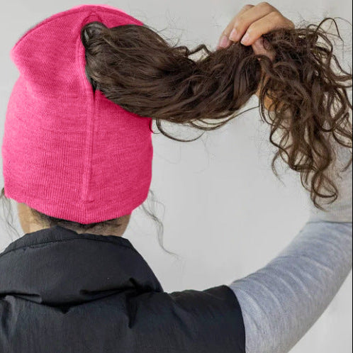Person wearing a pink beanie and black vest, adjusting hair against a plain background