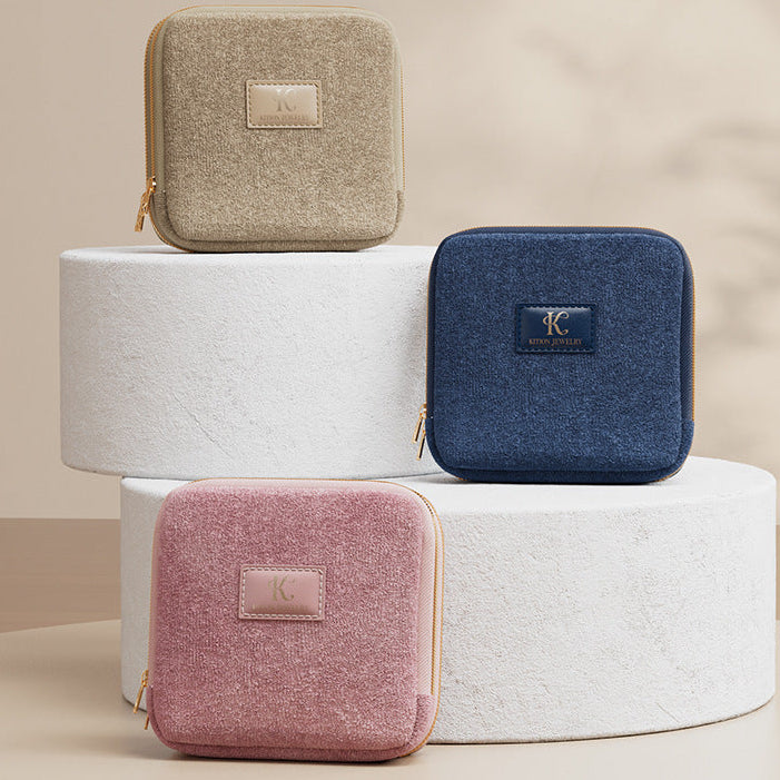 Three square pouches in beige, blue, and pink on a white cylindrical stand with a beige background.
