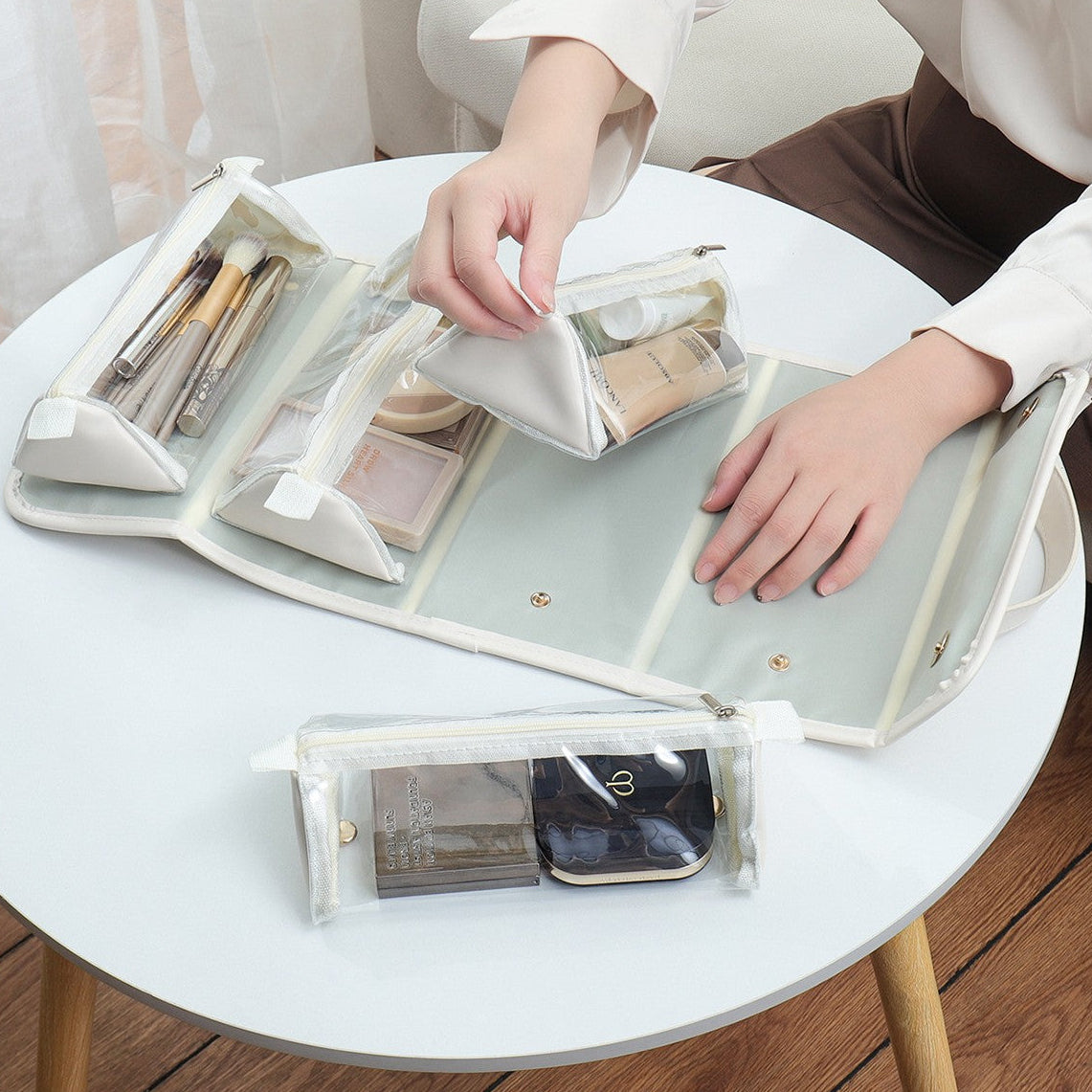 Person organizing makeup items in a transparent travel case with detachable bags on a white table.