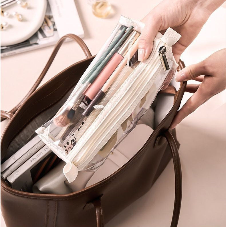 A clear plastic storage pouch containing makeup brushes and pens, being inserted into a brown handbag.