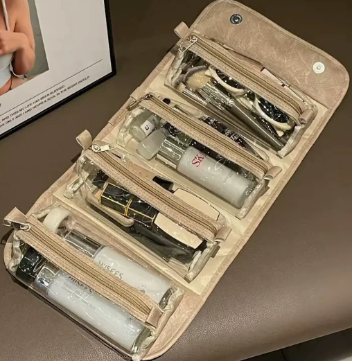 A taupe faux-leather travel makeup bag with clear zippered compartments, shown open and filled with cosmetics and skincare items on a brown leather surface.
